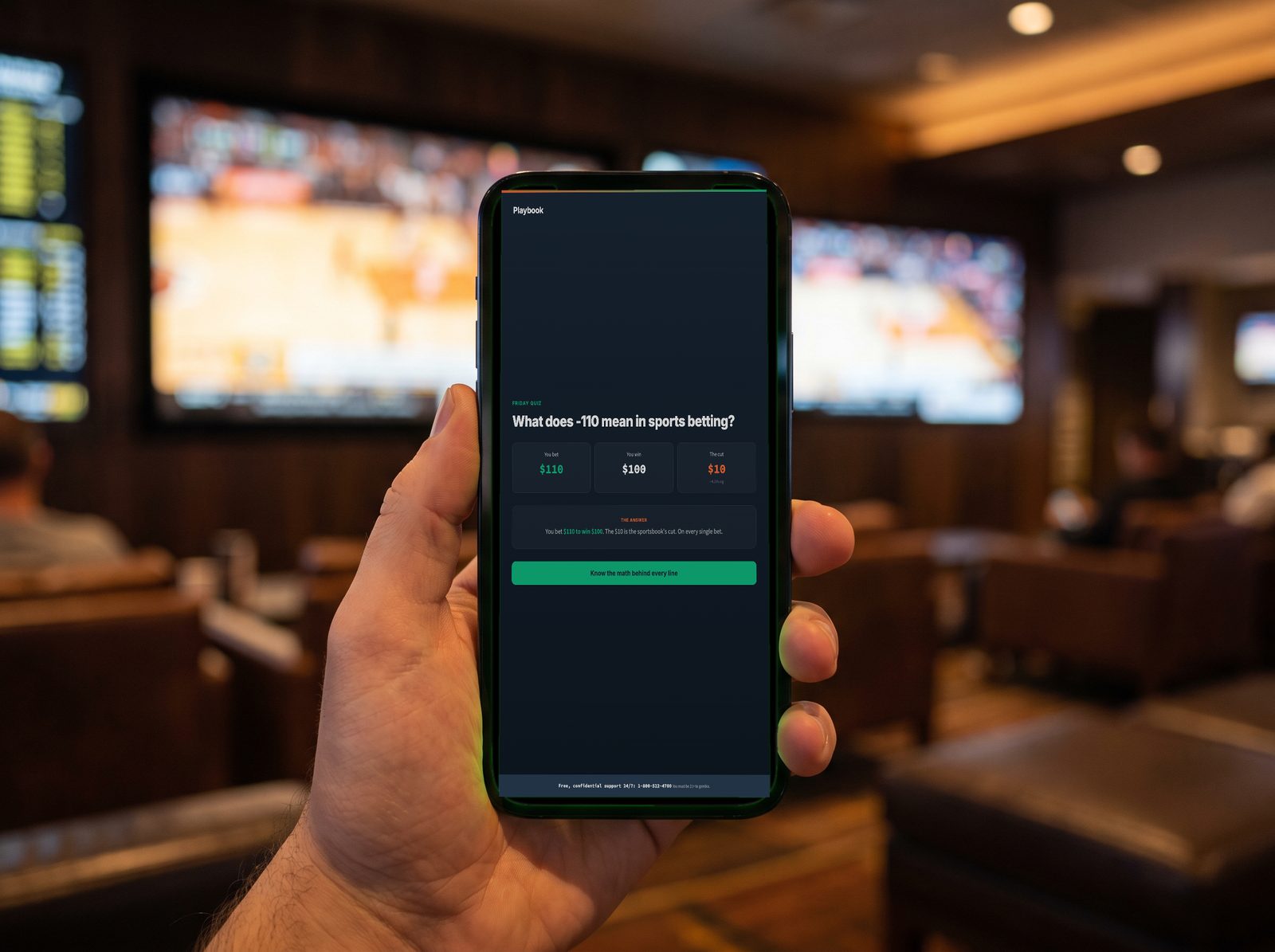 A phone at the book showing the Playbook story card “What does -110 mean in sports betting?” with the payout broken out.
